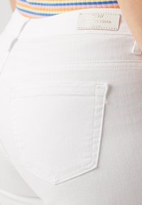 Los pantalones vaqueros de mezclilla blanca cuentan con un bolsillo trasero que tiene detalles de costura y una etiqueta de marca en la parte superior. La tela tiene una apariencia suave y texturizada.