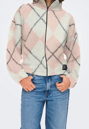 Person trägt eine weiß-rosa karierte Fleecejacke mit schwarzem Reißverschluss, die Hände in den Taschen einer blauen Jeans, stehend vor weißem Hintergrund.