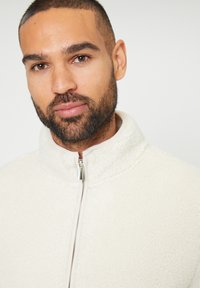 Giacca in pile bianca con colletto alto e chiusura frontale con zip. Caratterizzata da una texture morbida e cuciture sottili; design pulito e minimalista.