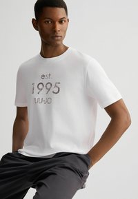 Witte katoenen t-shirt met een gestructureerde, metallic print met "est. 1995 LIUJO" gecentreerd aan de voorkant; ronde hals en korte mouwen.