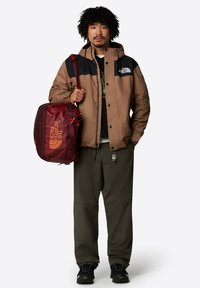 Brązowa i czarna wodoodporna kurtka noszona na zielonej koszuli, w zestawieniu z oliwkowymi spodniami i czarnymi butami. Nosi bordo plecak z pomarańczowym logo.