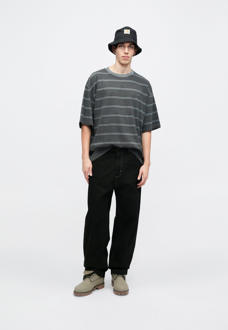 Gestreiftes schwarz-weißes Oversized-T-Shirt, schwarze lockere Hose und grüne Wildlederstiefel mit Gummisohlen. Schwarzer Bucket Hat mit einem Logo-Patch.