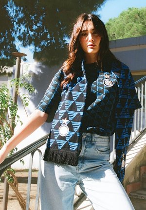 Vrouw die buiten staat, draagt een blauw-zwart patroon sjaal en bijpassend overhemd, gecombineerd met lichtblauwe spijkerbroek, nabij een metalen reling en groen.