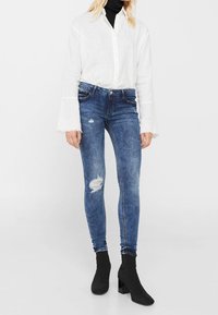 Pantalones vaqueros ajustados de mezclilla azul con roturas desgastadas, combinados con una camisa blanca abotonada y botines negros. Diseño informal y entallado.