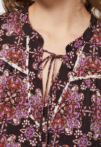 Chemisier à fleurs sombres avec un fermoir à nouer à l'avant, présentant des motifs complexes en rouge, violet et blanc, le tout en tissu léger et texturé.