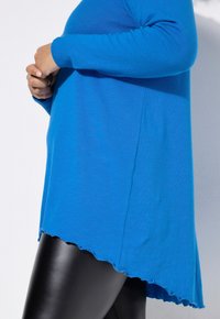 Blaues Langarmoberteil mit abgerundetem Saum und gewelltem Rand, aus weichem Material. Kombiniert mit glänzenden schwarzen Leggings.