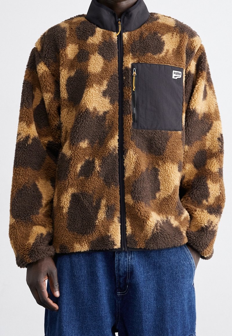 Hombre con chaqueta de forro polar marrón y marrón oscuro con estampado, bolsillo en el pecho con cremallera negra y pantalones vaqueros azules.