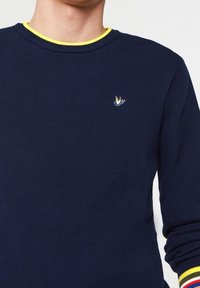 Marinblått texturerat tröja med en gul halsringning. Har en liten broderad fågel-logotyp på bröstet och randiga muddar med röda och gula accenter.