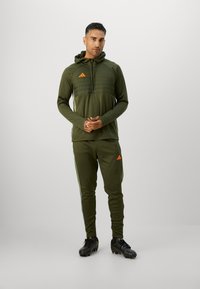 Felpa verde oliva con zip frontale e pannelli testurizzati, abbinata a pantaloni tapered coordinati con righe laterali e loghi. Calzature nere.