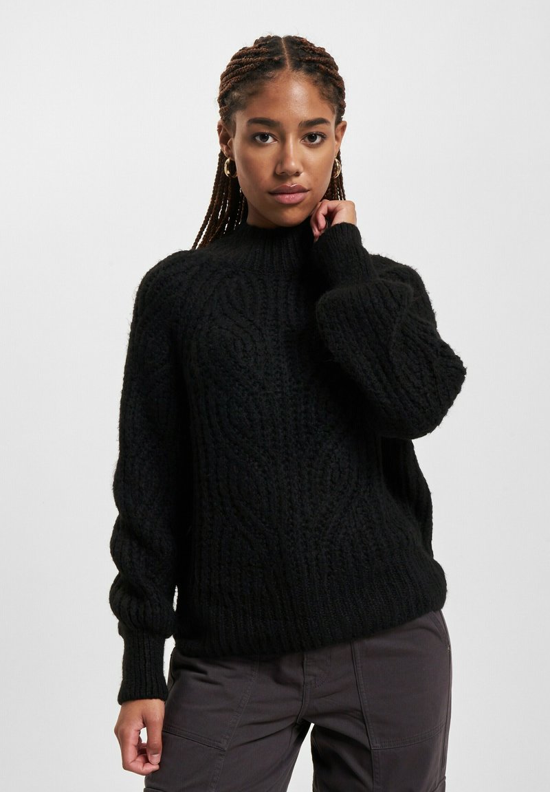 Schwarzer Strickpullover mit hohem Kragen, der ein strukturiertes, Zopfstrickmuster und lockere Ärmel hat. Getragen mit grauen Cargo-Hosen.