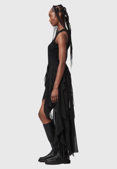 Femme aux longues tresses portant une robe noire asymétrique sans manches et des bottes noires montantes jusqu'aux genoux, debout de profil sur un fond gris.
