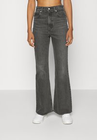 Jeans de mezclilla negra acampanada con cintura alta, tejido resistente, diseño clásico de cinco bolsillos y costuras en contraste, combinados con zapatillas blancas.