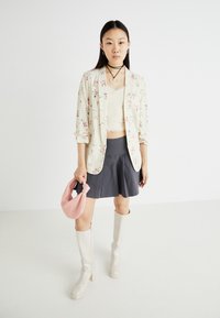 Blazer crème à motifs floraux, jupe grise ajustée, crop top beige, sac à main rose, bottes blanches à hauteur de genoux. Fond neutre.
