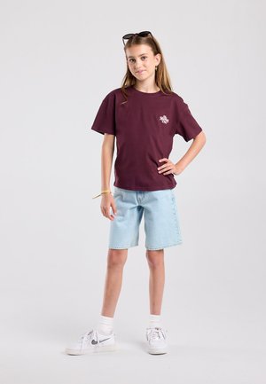 Tienermeisje staat met hand op heup, draagt een bordeauxrood T-shirt, lichtblauwe denim shorts, witte sneakers en een zonnebril op haar hoofd.