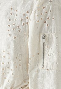 Närbild av vitt tyg med ögla broderi och en silverfärgad dragkedja på en ärmlomme.