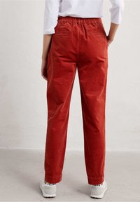 Pantalon en velours côtelé de couleur rouille avec une ceinture élastique, coupe droite et poches arrière, porté avec des baskets blanches.