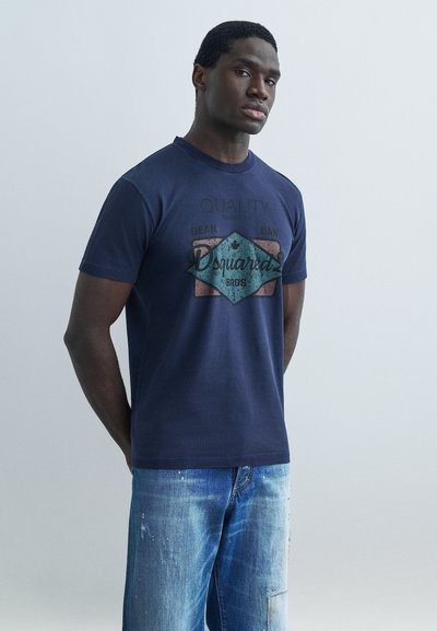Mann stående med hendene bak ryggen, iført navyfarget t-skjorte med grafisk trykk og blå jeans med slitt utseende mot en ensfarget lys grå bakgrunn.
