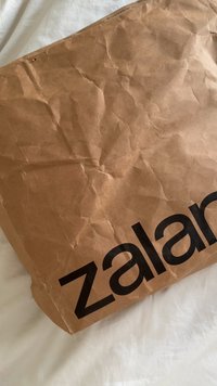 Saco de papel castanho amassado com o logótipo preto "zalando" impresso na lateral, repousando sobre uma superfície branca.