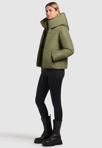 Olijfgroene pufferjas met een hoge kraag, ritssluiting en gewatteerd design. Gecombineerd met zwarte legging en kniehoog, veterschoenen.