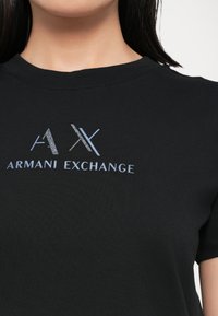 Juodas marškinėliai, pagaminti iš glotnaus audinio, su metalizuotu pilku ir mėlynu "AX" logotipu bei "ARMANI EXCHANGE" tekstu priekyje. Apvalus iškirpimas.