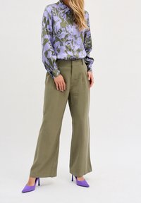 Blusa a maniche lunghe con motivo floreale lilla e verde, abbinata a pantaloni larghi a vita alta verde oliva e tacchi alti viola.