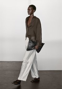 Brun gennemsigtig skjorte med knapper, lyse beige linnedbukser, sort tekstureret clutch og sorte sandaler med multicolore detaljer.