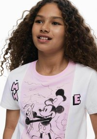 Hvid og lyserød t-shirt med korte ærmer, der viser en sort-hvid skitse af Mickey Mouse, med fed grafisk skrifttype på venstre side.