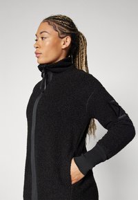 Black fleece jacket with a zip-up front and high collar. Features textured fabric, elastic cuffs, and side pockets.