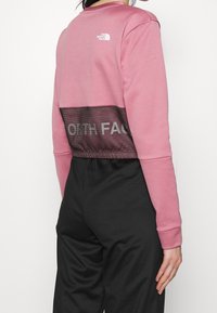 Osoba w różowej koszuli z długim rękawem z logo The North Face i czarnych spodniach, widziana z tyłu na białym tle.