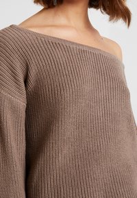 Hnědý pletený svetr bez ramene s texturovaným žebrováním, širokými rukávy a volným střihem, vyrobený z měkkého a teplého materiálu.