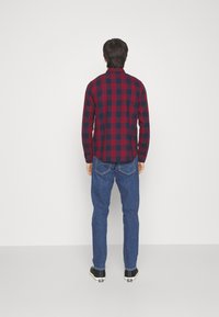Rød og marineblå ternet skjorte, lange ærmer, tætsiddende pasform, båret med blå denimjeans og sorte sneakers. Bagfra ses skjortens buede kant.