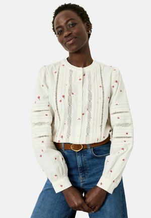 Weiße Hemdbluse mit roter Blumenstickerei, Spitzendetails und plissierten Akzenten; kombiniert mit blauen Jeans und braunem Gürtel.