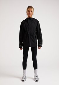 Svart lättviktsjacka med huva, elastisk midja, sidofickor och dragkedja. Bärs med svarta leggings och sneakers.