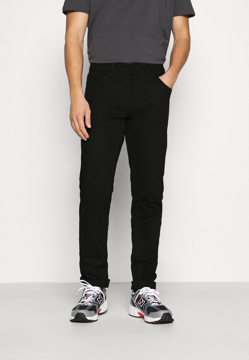 Homme portant un pantalon noir, un t-shirt gris et des baskets New Balance noires, blanches et rouges, debout avec une main dans la poche.