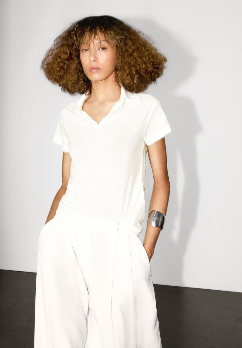 Bílá polokošile vyrobená z měkké tkaniny s límečkem, kombinovaná s volnými bílými kalhotami. Na ruce má hladký stříbrný náramek.