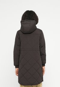 Person står med ryggen til, iført en mørk, kviltet lang jakke med hætte og beige bukser mod en ensfarvet hvid baggrund.