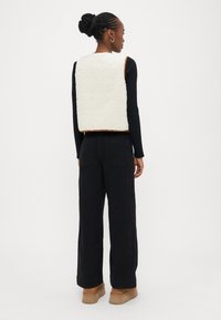 Hvit, myk fleecevest med rund hals, brun kant, brukt over en svart langermet skjorte og kombinert med svarte bukser med vide ben.