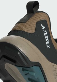 Buty Terrex mają teksturowaną cholewkę w brązowo-czarnym kolorze, z formowaną beżową podeszwą oraz charakterystycznym turkusowym akcentem na podeszwie zewnętrznej.