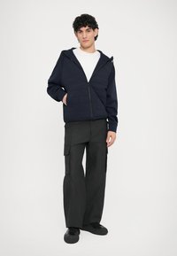 Μπλε-σκούρο φούτερ με φερμουάρ και επένδυση καπιτονέ, φορεμένο πάνω από λευκό πουκάμισο, σε συνδυασμό με χαλαρό μαύρο cargo παντελόνι με πλαϊνές τσέπες. Μαύρα αθλητικά παπούτσια.