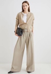 Cardigan lavorato a maglia beige con bottoni, abbinato a pantaloni beige chiaro a vita alta e gamba larga, e una borsa a tracolla nera, posizionato su uno sfondo neutro.