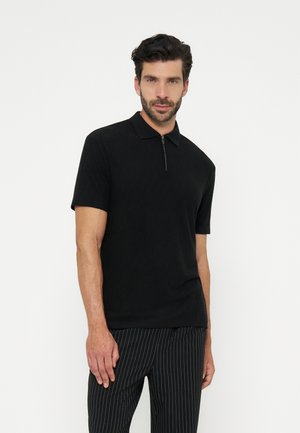 Homme portant un polo noir à manches courtes avec une fermeture éclair sur le devant et un pantalon noir à fines rayures, debout devant un fond blanc uni.