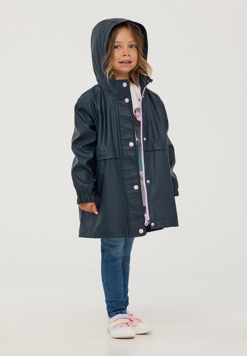 Impermeabile blu navy con cappuccio, bottoni a pressione rosa, polsini elastici e una cerniera frontale sopra una camicia bianca a fantasia colorata.