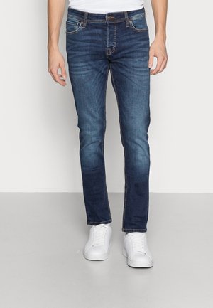 Jean bleu foncé avec une coupe slim, comportant des poches, des détails brodés et une texture vieillie. Assortis à des baskets blanches à lacets.