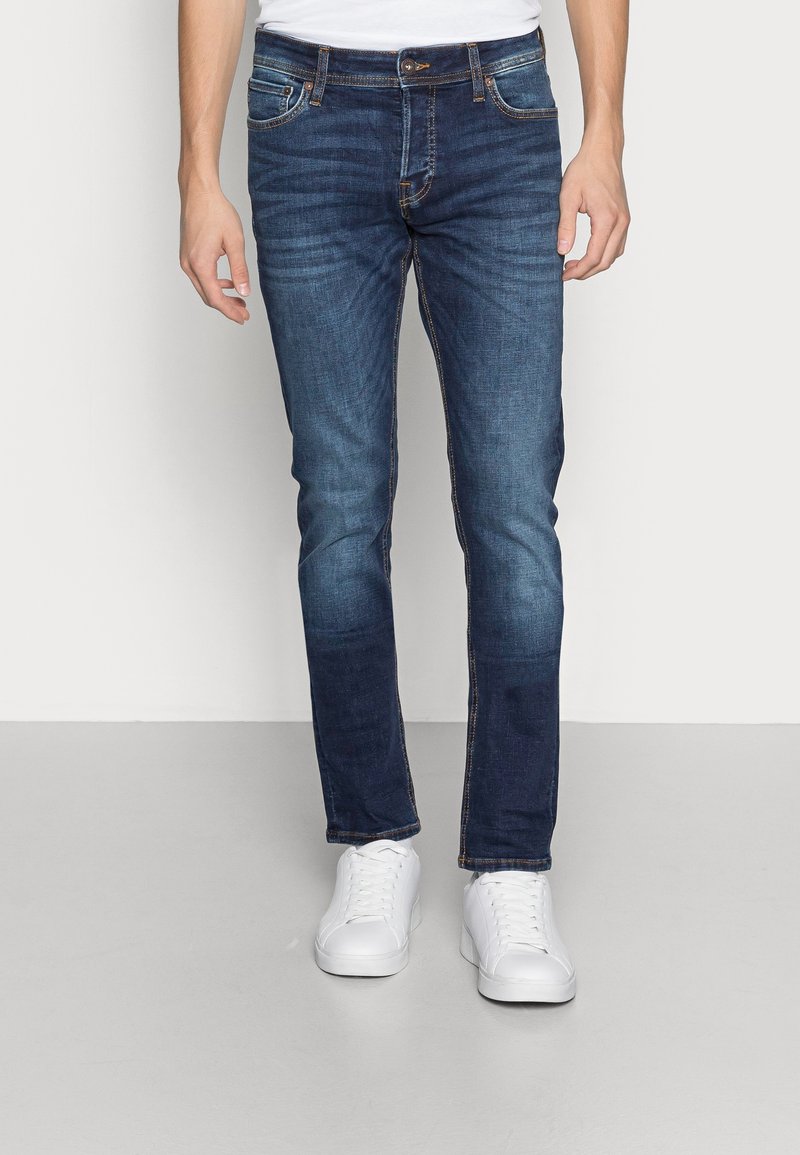 Jean bleu foncé avec une coupe slim, comportant des poches, des détails brodés et une texture vieillie. Assortis à des baskets blanches à lacets.