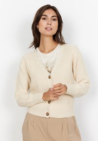 Beige knitted cardigan featuring a V-neck, long sleeves, and brown buttons. The texture is ribbed, with a fitted waistline.