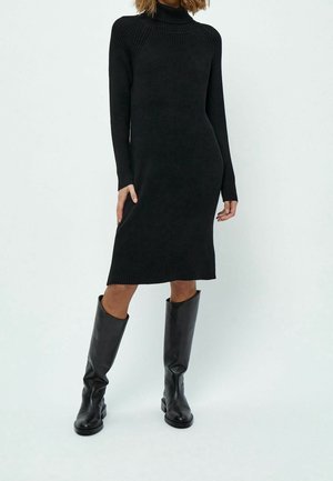 Femme portant une robe midi noire à col roulé côtelée, associée à des bottes en cuir noires hautes jusqu'aux genoux, debout devant un fond clair uni.