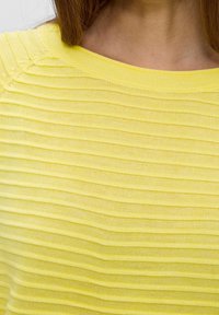 Gul stickad topp med ribbad textur, rundad halsringning och detaljerade sömmar. Lätt och andningsbart tyg med en mjuk finish.