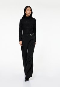Sort strikket bluse med lange ærmer og tekstureret mønster, parret med højtaljede, vide sorte bukser og et bredt sort bælte.
