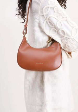 Sac à bandoulière en cuir marron avec des ferrures dorées porté par une personne portant un pull blanc texturé avec un motif floral sur la manche.