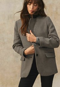 Jachetă cu un model houndstooth alb-negru, cu guler înalt și două buzunare frontale, având o textură fină și o croială ajustată.